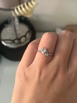 Silverring med hjärtformad sten - Säljer en snygg silverfärgad ring med en stor hjärtformad klar sten i mitten och mindre stenar längs bandet. Ringen har en elegant och romantisk design som verkligen sticker ut. De är inget problem med den, dock finns de ingen ”låda” som den brukar ligga i så därför säljer jag den lite billigare. 