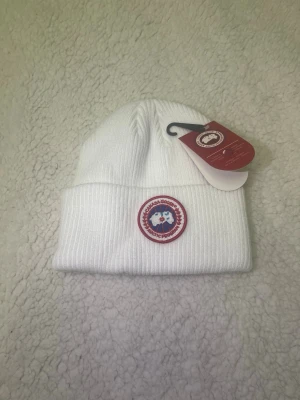 Vit ribbstickad mössa Canada Goose - Snygg vit ribbstickad mössa från Canada Goose med klassisk rund logotyp framtill. Mössan har uppvikt kant och är tillverkad i mjukt material som håller dig varm. Perfekt accessoar för kyliga dagar och en stilren detalj till din outfit.
