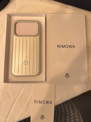Rimowa silver mobilskal i aluminium - Snyggt mobilskal från Rimowa i stilren silverfärg med ikoniska räfflade linjer. Tillverkat i aluminium och har en modern, minimalistisk design med Rimowa-loggan på baksidan. Perfekt för dig som vill ha ett exklusivt och tåligt skal till mobilen.