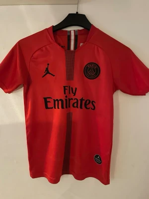 Röd PSG Neymar Jr matchtröja - Snygg röd Paris Saint-Germain matchtröja med Neymar Jr och nummer 10 på ryggen. Tröjan har Fly Emirates-tryck på framsidan, svarta detaljer och Jordan-logga. Perfekt för fotbollsfans som vill sticka ut på planen eller läktaren.