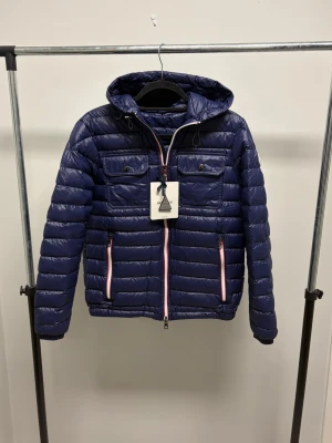 Moncler Jacka - Mörkblå jacka med luva från Moncler. Dragkedjan har en liten skada som kan göra så att den blir segare ibland över den lilla delen, annars nyskick. Tags tillkommer. Storlek 2 så sitter som en small/liten medium.