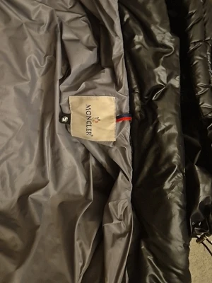 Svart dunjacka från Moncler - Säljer en svart dunjacka från Moncler med glansig finish och huva. Jackan har grått innerfoder, dragkedja och klassisk Moncler-logga på insidan. Perfekt för kalla vinterdagar och riktigt snygg streetstil.