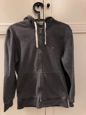 Mörkgrå hoodie Tommy Hilfiger - Mysig mörkgrå hoodie från Tommy Hilfiger med dragkedja, vit fleece på insidan och vita snören i huvan. Klassisk liten logga på bröstet och stora fickor framtill. Perfekt för chill dagar.