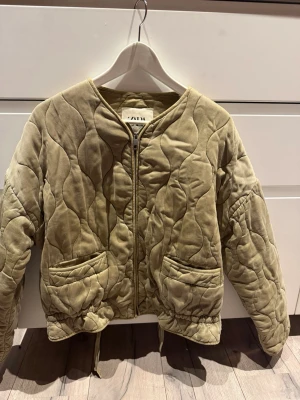 Beige quiltad cardiganjacka från Zara - Quiltad beige cardiganjacka från Zara med dragkedja framtill, två stora fickor och rund halsringning. Jackan har vågigt mönster i tyget och dragsko i nederkant för justerbar passform. Perfekt för lager på lager och en chill look.