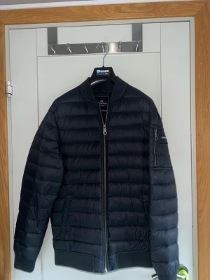 Mörkblå pufferjacka från Lexington - Snygg svart pufferjacka från Lexington med quiltad design och ribbade muddar. Jackan har dragkedja framtill, fickor på sidorna och en extra ficka med dragkedja på ärmen. Perfekt för kalla dagar och riktigt stilren look.