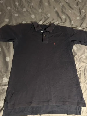 Mörkblå pikétröja från Ralph Lauren - Klassisk mörkblå pikétröja från Ralph Lauren med krage, knappar och den ikoniska broderade loggan i rött på bröstet. Tröjan är tillverkad i bomull och har korta ärmar, perfekt för en stilren och avslappnad look.