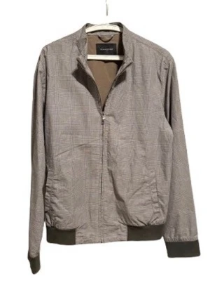 Grå bomberjacka med rutmönster - Snygg grå bomberjacka från Zara Man med diskret rutmönster. Jackan har dragkedja framtill, ribbade muddar vid ärmslut och nederkant samt ståkrage. Tillverkad i ett lätt material som passar perfekt till lager på lager. Enkel och stilren design.