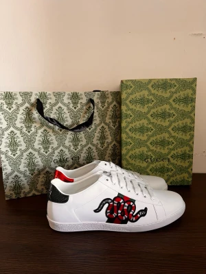 Gucci skor - Snygga vita sneakers från Gucci med ikonisk broderad orm i rött, svart och vitt på sidan. Skorna har klassisk snörning, gröna och röda detaljer på hälen och är tillverkade i slätt skinn. Perfekta för dig som vill sticka ut med lyxig streetstyle. Pris kan diskuteras vid snabb affär 