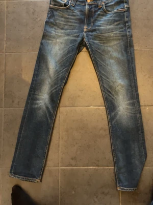 Blå jeans från Nudie Jeans - Säljer ett par klassiska blå jeans från Nudie Jeans med snygg slitning och raka ben. Jeansen har fem fickor, orange sömmar och Nudie-logga på bakfickorna. Perfekta för en avslappnad och trendig look.