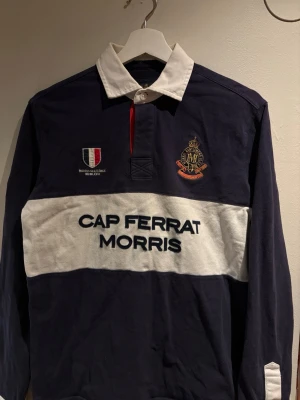 Morris långärmad rugbytröja navy - Snygg långärmad rugbytröja från Morris i marinblått med vit krage och bred vit rand över bröstet. Broderade detaljer och texten 'CAP FERRAT MORRIS' framtill. Klassisk preppy stil med brodyr och kontrastfärgad knappslå.
