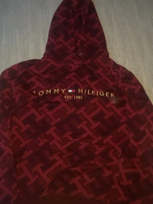 Vinröd hoodie från Tommy Hilfiger - Säljer en vinröd hoodie från Tommy Hilfiger med stort H-mönster över hela tröjan och gul broderad logga samt flagga på bröstet. Hoodien har huva med dragsko och en stor magficka. Perfekt för dig som gillar streetwear och snygga detaljer. Storlek XL men sitter som L