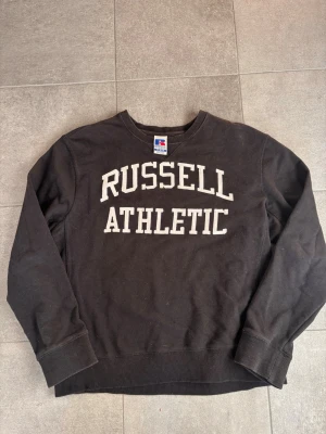 Svart sweatshirt från Russell Athletic - Klassisk svart sweatshirt från Russell Athletic med vit broderad text på bröstet. Tröjan har rund halsringning, ribbade muddar vid ärmslut och nederkant samt en avslappnad passform. Perfekt för en sportig och avslappnad stil.