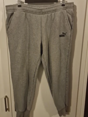 Gråa mjukisbyxor från Puma - Mysiga gråa mjukisbyxor från Puma med resår i midjan och muddar vid bensluten. Byxorna har två sidofickor och Puma-logga tryckt på vänster lår. Perfekta för chill eller träning.