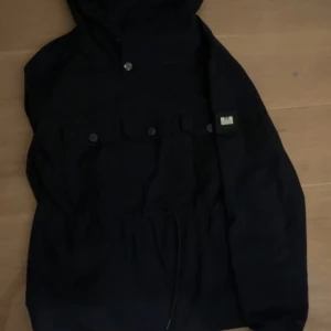 Weekend offender jacka  - Mörkblå jacka från weekend offender med huva och snören i midjan. Jackan har knappstängning upptill, tre bröstfickor med lock och en liten logotyp på vänster sida. Perfekt för blåsiga dagar och enkel att matcha till olika outfits.