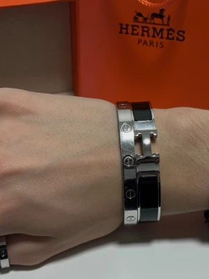 Svart och silver armband - Stilrent armband i silverfärgad metall med svarta detaljer. Armbandet har den ikoniska H-formade spännet och blanka ytor som ger en lyxig känsla. Perfekt accessoar för att lyfta din outfit med en exklusiv touch.