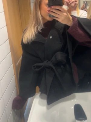 Svart kappa - Snygg svart kappa med bred krage och knytskärp i midjan. Kappan har en lös passform och är ifrån Gina Tricot.. Stängs med knappar uptill och har en stilren look som passar både vardags och fest tillfällen! 