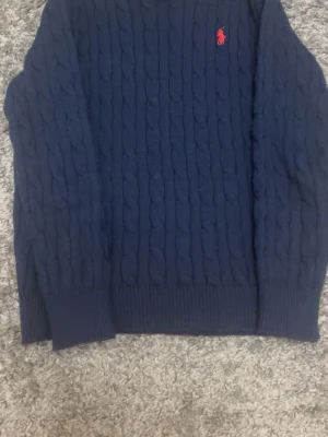 Mörkblå kabelstickad tröja Polo Ralph Lauren - Kabelstickad mörkblå tröja från Polo Ralph Lauren med klassisk röd logga broderad på bröstet. Tröjan har rund halsringning och långa ärmar, perfekt för lager-på-lager-stil. Materialet är mjukt och värmande stickat. Passar också som XS