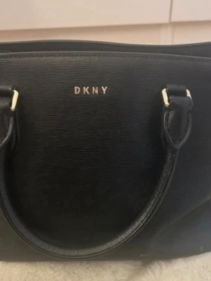 Svart handväska från DKNY? - Snygg svart handväska från DKNY med guldfärgade detaljer och två rejäla handtag. Väskan är tillverkad i strukturerat konstläder och har DKNY-logga framtill. Insidan är fodrad med svart tyg och har en innerficka med dragkedja.                               Jag vet inte om den är äkta, därav priset.