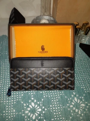 Svart Goyard plånbok med mönster - Snygg svart plånbok från Goyard med klassiskt grafiskt mönster i vitt och brunt. Plånboken har dragkedja runtom och är tillverkad i canvas med läderdetaljer. Perfekt för dig som gillar exklusiva accessoarer och vill ha något som sticker ut.