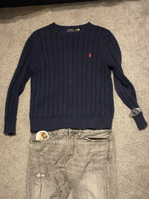 Mörkblå kabelstickad tröja Polo Ralph Lauren - Säljer en mörkblå kabelstickad tröja från Polo Ralph Lauren med den klassiska röda loggan broderad på bröstet. Tröjan har rund halsringning och långa ärmar, perfekt för dig som gillar stilrena och tidlösa plagg.