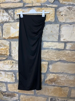 Svart långkjol med rynkdetalj - Stilren svart långkjol med snygg rynkdetalj på ena sidan. Kjolen har en rak passform och är tillverkad i ett mjukt, stretchigt material som ger en bekväm känsla. Perfekt att matcha med en crop top eller en oversized tröja.