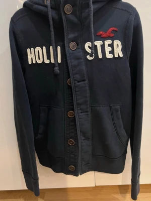 Marinblå Hollister cardigan  - Mysig marinblå cardiganjacka från Hollister med huva och knappar framtill. Stor broderad logga och text på bröstet, två stora fickor och dragsko i huvan. Perfekt för lager på lager och chill stil. Denna är väldigt eftertraktad och är i väldigt bra skick 9/10 den är perfekt nu till hösten då den är ganska varm. Kom privat vid frågor eller andra funderingar//🤝