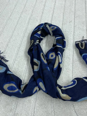 Mönstrad blå halsduk med fransar - Snygg halsduk i mörkblått med abstrakt mönster i beige, ljusblått och svart. Tunn och lätt i materialet, perfekt att slänga över axlarna. Fransar i ändarna ger en avslappnad vibe. Passar dig som gillar färg och mönster.