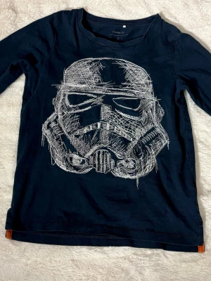 Mörkblå långärmad topp Name It - Mörkblå långärmad topp från Name It med vitt grafiskt tryck framtill. Toppen har rund halsringning och kontrastsömmar i orange vid nacken. Perfekt för en avslappnad och cool stil.