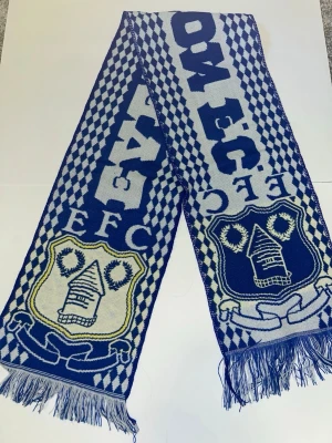 Blåvit Everton FC halsduk - Supporterhalsduk i blått och vitt med Everton FC:s klubbmärke och texten 'EFC'. Halsduken har rutmönster längs sidorna och fransar i ändarna. Perfekt för att visa ditt lagstöd på matchen eller i vardagen.