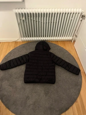 Svart pufferjacka från Lyle & Scott - Svart pufferjacka med huva från Lyle & Scott. Jackan har ett quiltat mönster, dragkedja framtill och två fickor. På bröstet sitter den klassiska gula loggan. Perfekt för kalla dagar och riktigt skön att bära.