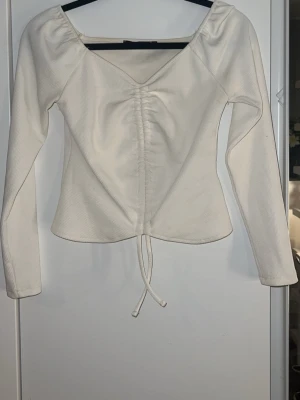 Vit ribbad långärmad topp med snörning - Trendig vit långärmad topp med ribbad struktur och snygg v-ringning. Toppen har snörning framtill som ger en cool detalj och croppad känsla. Perfekt att styla med jeans eller kjol för en fräsch look.
