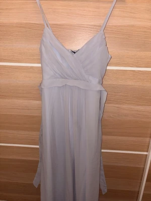 Ljusblå långklänning med omlottopp - Snygg ljusblå långklänning med omlott över bysten och smala axelband. Klänningen har ett elegant fall och markerad midja, perfekt för speciella tillfällen. Materialet är lätt och luftigt. Justerbara band och dragkedja på sidan. Aldrig använd. Ingen prislapp dock.