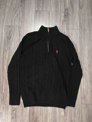 Svart kabelstickad tröja Polo Ralph Lauren - Svart kabelstickad tröja från Polo Ralph Lauren med halv dragkedja och hög krage. Tröjan har ett rött broderat logomärke på bröstet och ribbstickade muddar. Perfekt för en stilren och klassisk look.