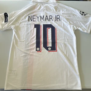 PSG Neymar Jr 10 vit matchtröja Nike - Säljer en Paris Saint-Germain fotbollströja med Neymar Jr och nummer 10 på ryggen. Två små prickar enligt bild 4 och 5, därav priset! Tröjan är vit med röda och marinblå detaljer, har korta ärmar, krage och Champions League-märke på ärmen. Materialet är lätt och andas, perfekt för fotboll eller supporterliv. Replika, endast handtvätt gäller!