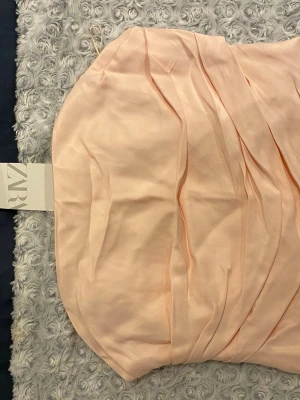 Ljusrosa axelbandslös klänning Zara - Söt axelbandslös klänning från Zara i ljusrosa nyans. Klänningen har draperade detaljer framtill och är tillverkad i ett slätt, glansigt material som ger en elegant känsla. Perfekt för dig som gillar en feminin och stilren look.