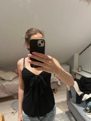 Svart linnetopp med knyt fram - Snygg svart linnetopp med smala axelband och knytning framtill. Toppen har en djup v-ringning och är figurnära med en croppad känsla. Perfekt att styla med jeans eller kjol för en trendig look.