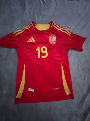 Spaniens landslagströja nr 19 Lamine Yamal Adidas - Röd fotbollströja från Adidas med gula detaljer, Spaniens landslagsemblem på bröstet och nummer 19. Namnet 'Lamine Yamal' står på ryggen. Tillverkad i funktionsmaterialet HEAT.RDY för bästa komfort under match eller träning.