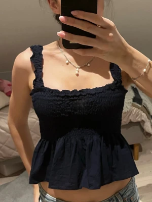 Mörkblå smockad linnetopp - Supersöt mörkblå linnetopp med breda axelband och smockad överdel. Toppen har volang nedtill och en lätt croppad passform som ger en avslappnad men ändå feminin vibe. Perfekt att styla med jeans eller kjol för en trendig look.