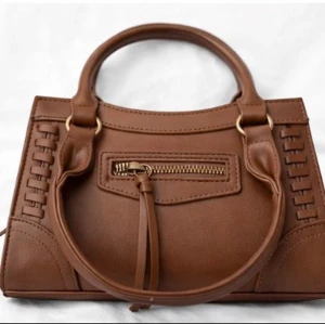 Brun handväska med gulddetaljer - Snygg brun handväska i konstläder med rundade handtag och guldfärgade metalldetaljer. Väskan har en dragkedjeficka framtill med dekorativ tofs och flätade detaljer längs sidorna. Perfekt accessoar för att lyfta din outfit. Skriv gärna för fler bilder✨✨