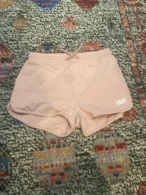Ljusrosa sportshorts från WA - Snygga ljusrosa sportshorts från WA med elastisk midja och dragsko. De har en avslappnad passform och är tillverkade i ett lätt och mjukt material, perfekt för träning eller chill. Diskret logga i vitt på ena benet.