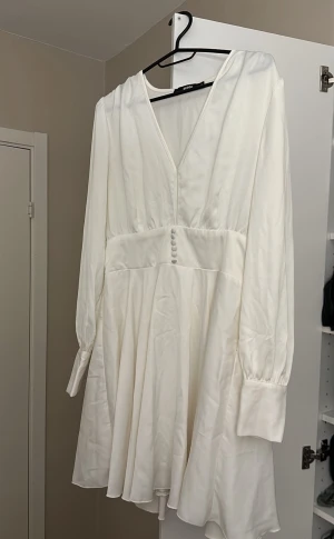 Vit omlottklänning från Bik Bok - Supersöt vit kortklänning från Bik Bok med v-ringning, långa ärmar och knappar framtill i midjan. Klänningen har omlottkänsla och är tillverkad i ett glansigt, mjukt material som ger en lyxig känsla.