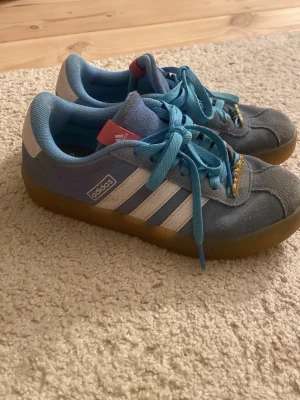 Blå Adidas sneakers med vita ränder - Säljer ett par klassiska Adidas sneakers i blått med vita ränder och detaljer. Skorna har ljusblå skosnören, gummisula och ovandel i mocka. Perfekta för dig som gillar retrostil och vill ha något bekvämt och snyggt till vardags.