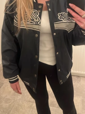 Svart vindjacka med vita detaljer - Snygg svart vindjacka i sportig modell med vita ränder och broderade detaljer på bröstet. Jackan har tryckknappar framtill, ribbade muddar och krage med vita ränder. Materialet är glansigt och tunt, perfekt för lager-på-lager. Passar dig som gillar streetwear och avslappnad stil. Köpt på Beyond retro i Zinkensdamm, ordinarie pris 800kr