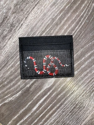 Svart korthållare med orm - Säljer en svart korthållare i skinn med GG-mönster på baksidan och en cool röd, vit och svart orm på framsidan. Perfekt för kort och ID, stilren och lyxig design som sticker ut i fickan.