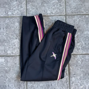 Axel arigato byxor  - Svarta mjukisbyxor från Axel arigato  likande Palm angels med lila, beige och vinröda ränder längs sidorna. Broderad fjäril på ena benet. Byxorna har resår i midjan och sidofickor. Perfekta för en avslappnad och trendig stil.