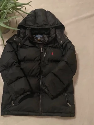 Svart pufferjacka från Polo Ralph Lauren - Snygg svart pufferjacka från Polo Ralph Lauren med huva och dragkedja framtill. Jackan har två sidofickor med dragkedja och den klassiska röda loggan broderad på bröstet. Perfekt för kalla dagar och riktigt stilren.