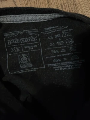 Svart Patagonia t-shirt med tryck XS - Svart t-shirt från Patagonia i regular fit med coolt bergsmotiv i blått, beige och vitt på bröstet. Tillverkad av 55% återvunnen polyester och 45% återvunnen bomull. Perfekt för dig som gillar stilren design med en twist.