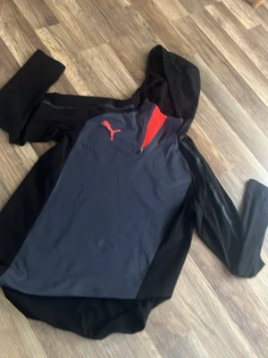 Svart och mörkgrå Puma tränings tröja - Snygg tränings tröja  från Puma i svart och mörkgrått med halv dragkedja och röd logga på bröstet. Insidan av huvan har en röd detalj som ger extra stil. Tillverkad i mjukt material, perfekt för en sportig och avslappnad look.