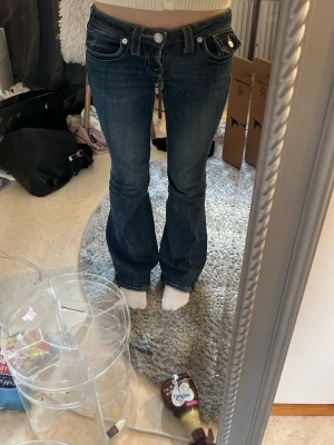 Mörkblå bootcut jeans True Religion - Säljer ett par mörkblå bootcut jeans från True Religion med kontrastsömmar och ikoniska fickdetaljer bak. Jeansen har låg midja, klassisk femficksdesign och silverfärgade knappar. Lite slitna nere vid fötterna men inget som man lägger märke till 