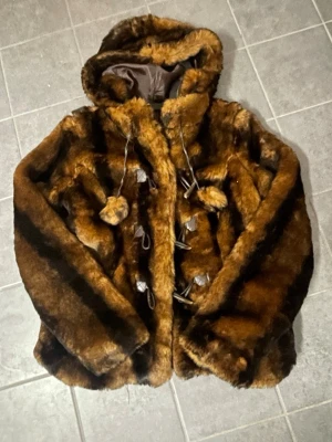 Brun pälsjacka med huva - Säljer en fluffig brun pälsjacka med huva och dekorativa tofsar. Jackan har stora knappar framtill och ett mysigt foder. Perfekt för dig som vill ha en varm och snygg jacka med lite extra attityd.